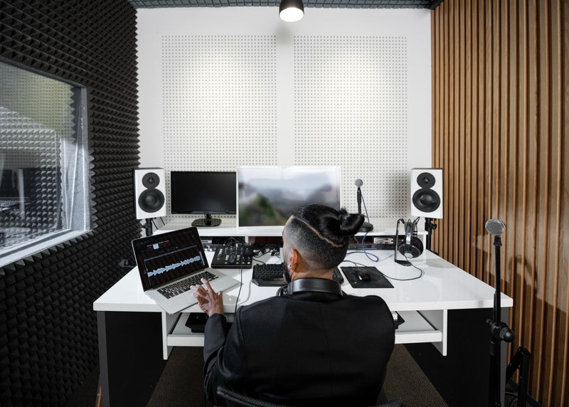 Man in acoustically treated DJ studio using laptop and audio equipment, surrounded by foam panels and studio monitors